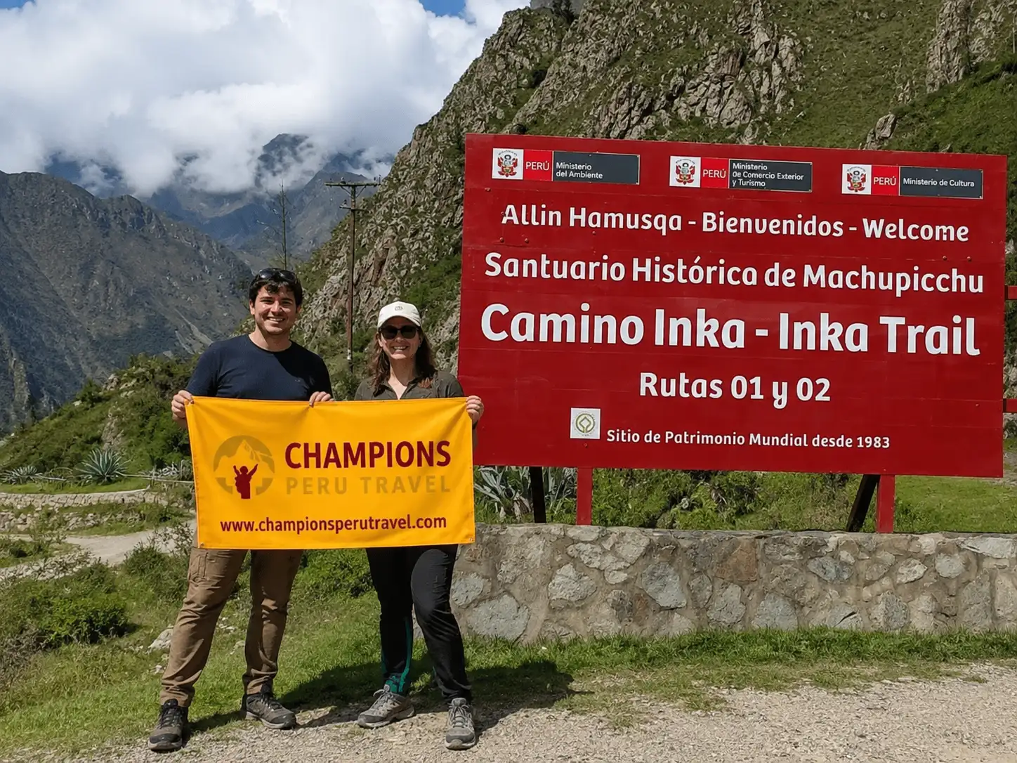 Two travelers holding a Champions Peru Travel banner beside the official Inca Trail entrance sign near Machu Picchu.