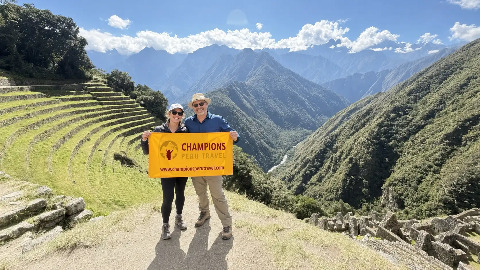 Two travelers holding a Champions Peru Travel banner beside Inca terraces and mountain scenery along the trail in Peru.